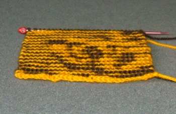 A small knitted swatch of the pattern that is designed. The swatch is knit with orange and black yarn and is viewed at an angle, revealing a portion of the Tiger's face.
