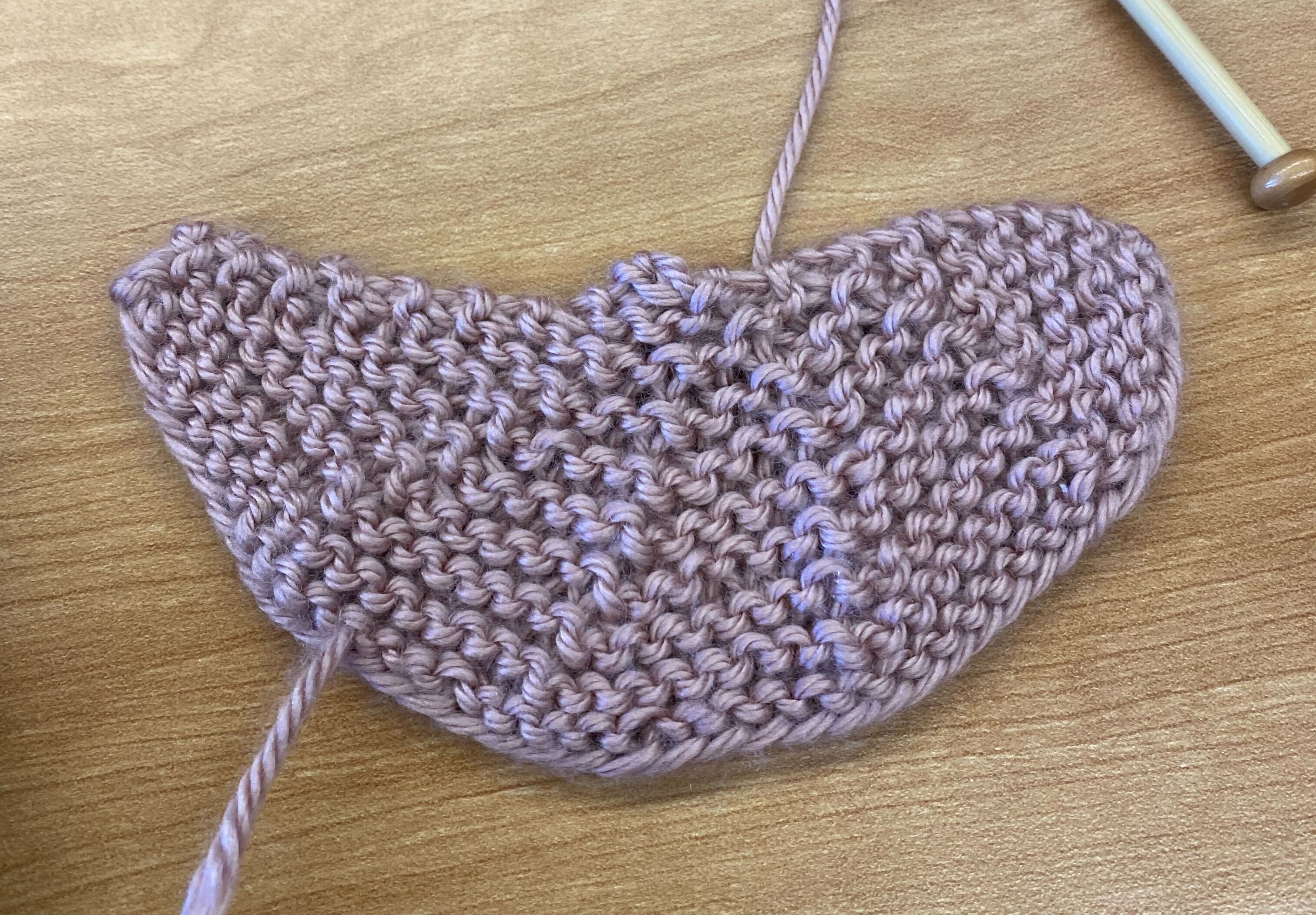 A small pink knit flat piece, about a semicircle in shape. It is divided into five different roughly triangular sections where the stitches are all parallel within the sections, but the angle from horizontal differs between sections. The angle starts at about -70 at the left side, then -30, then 15. 60, and 90 roughly.