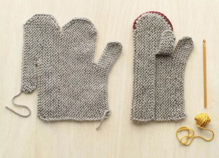 Diagram showing two mittens side-by-side in various stages of completion. The left mitten is a fully flat knit shape that has not yet been sewn together. It almost resembles the shape you would get if you cut a mitten along the fingertips and thumb and unfolded it, except on one side, the thumb flap is ponting down instead of up. On the right, the mitten has been folded over and the fingertips have been sewn. The thumb flap that was previously facing down has been flipped up so the thumb flaps can be sewn together.