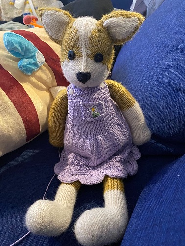 A hand-knit stuffed corgi on a blue couch that's somewhat anthrophomorphic, akin to a teddy bear. It is wearing a lavender pinafore dress, also knitted, with a leaf scallop edge.