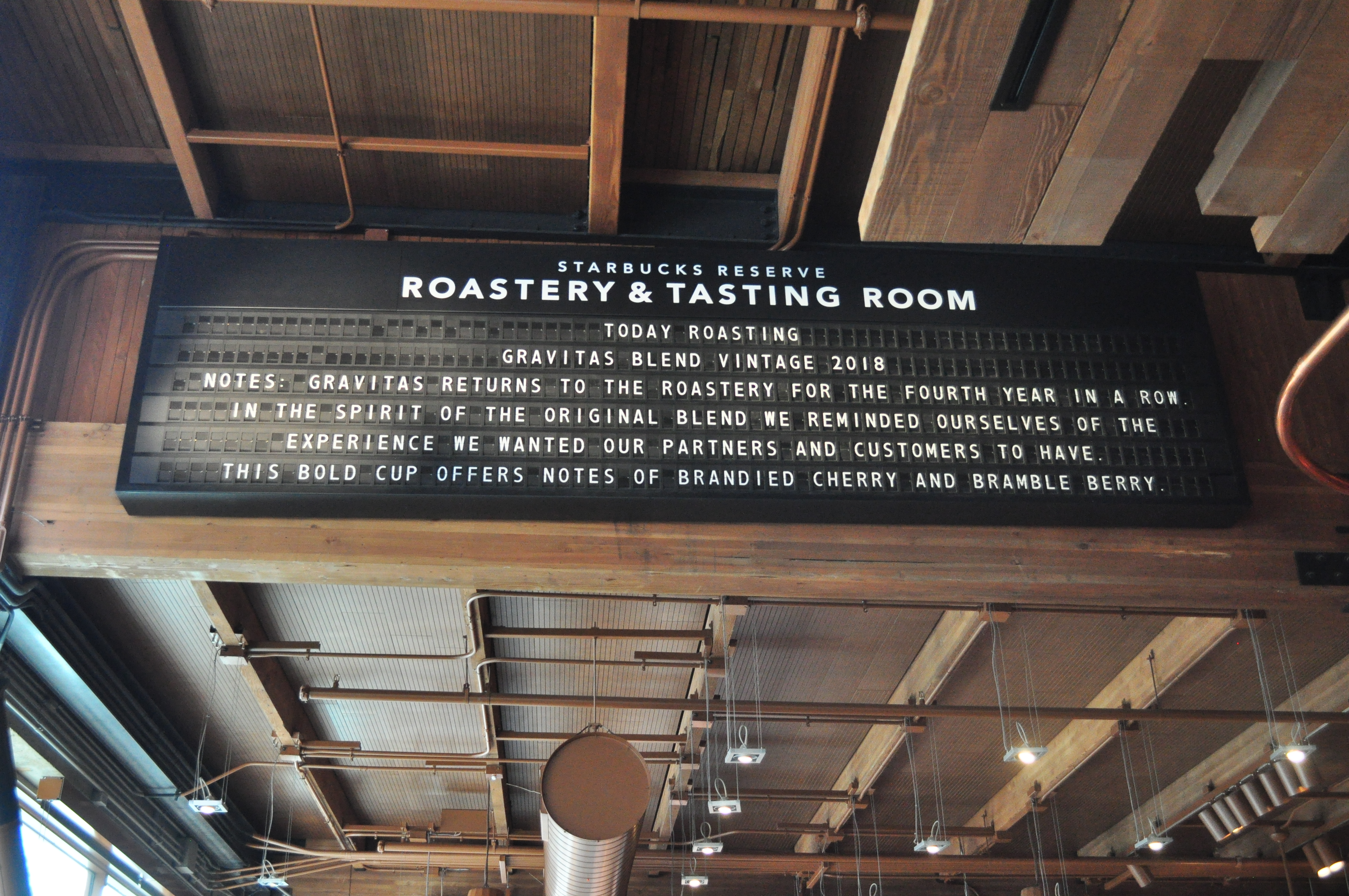 Large black split-flap sign with white lettering, with the words "Starbucks Reserve Roastery & Tasting Room" printed above the mechanism. The board is mounted near the ceiling on a wood partition. The text displayed by the split flap reads (backslashes denote line breaks): Today Roasting \ Gravitas Blend vintage 2018 \ Notes: Gravitas returns to the roastery for the fourth year in a row \ in the spirit of the original blend we reminded ourselves of the \ experience we wanted our partners and customers to have. \ This bold cup offers notes of brandied cherry and bramble berry."